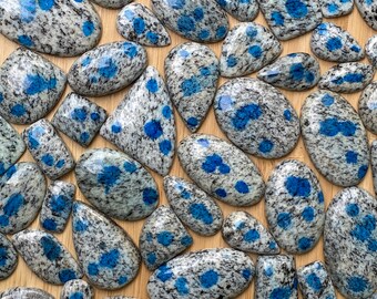 May include: A collection of polished K2 stone cabochons in various shapes, including oval, teardrop, and rectangular. The stones feature a speckled gray and white base with vibrant blue spots, arranged on a light brown wooden surface.