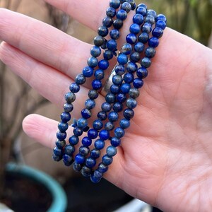 May include: A strand of blue lapis lazuli beads, with a speckled pattern, is held in a hand. The beads are round and uniform in size.