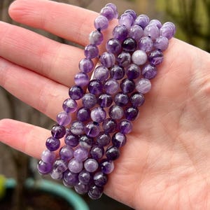 May include: A collection of amethyst bead bracelets. The beads are a mix of light and dark purple shades. The bracelets are strung on a dark cord. The bracelets are held in a hand.