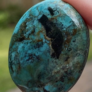 May include: A smooth, oval-shaped turquoise stone with a dark blue-green colour and black and brown matrix. The stone has a polished finish.