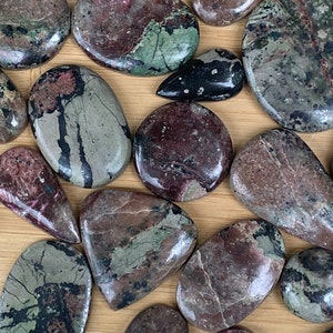 May include: Assortment of polished stones in various shapes, including ovals, teardrops, and irregular forms. The stones display a mix of colors, such as deep burgundy, muted green, and gray, with natural veining patterns. The collection is arranged on a light brown surface.