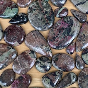 May include: A collection of polished gemstones in various shapes and sizes. The stones display a mix of colors, including deep reds, greens, and blacks, with unique patterns and textures. The gemstones are arranged on a light brown surface.