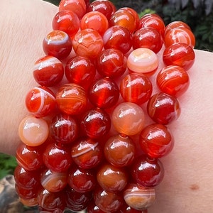Puede incluir: Un brazalete de piedra de cornalina con cuentas rojas y blancas con bandas. El brazalete está hecho de piedras de cornalina naturales y mide aproximadamente 20 cm de largo.