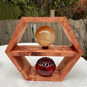 May include: A hexagonal wooden shelf with two shelves, displaying a light brown sphere on the top shelf and a red sphere with a decorative pattern on the bottom shelf.