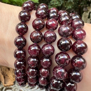 May include: A set of four garnet gemstone bracelets with dark red beads. The bracelets are stacked on a person's wrist.