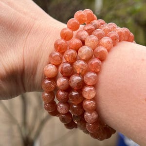 Puede incluir: Varios brazaletes de cuentas de color naranja rojizo. Las cuentas redondas tienen un aspecto moteado. Los brazaletes se llevan en la muñeca. El fondo está borroso, con toques de follaje verde.