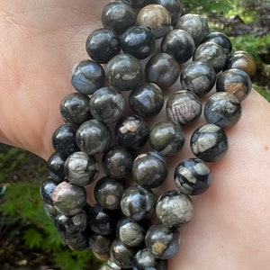 May include: A stack of black and grey gemstone bracelets with a speckled pattern. The bracelets are made of round beads and are worn on a person's wrist.