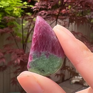 May include: A green and pink gemstone cabochon with a smooth, polished surface. The gemstone is held in a hand.