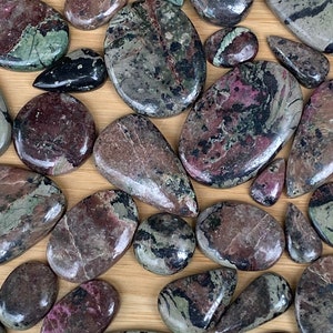 May include: A collection of polished stones in various shapes and sizes. The stones display a mix of colors, including deep reds, greens, and blacks, with intricate patterns. The stones are arranged on a light brown surface, showcasing their natural beauty.