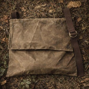 May include: A brown waxed canvas messenger bag with a fold-over flap closure. The bag features a dark brown adjustable fabric shoulder strap attached with a metal ring. The material has a textured, weathered appearance with visible creases.