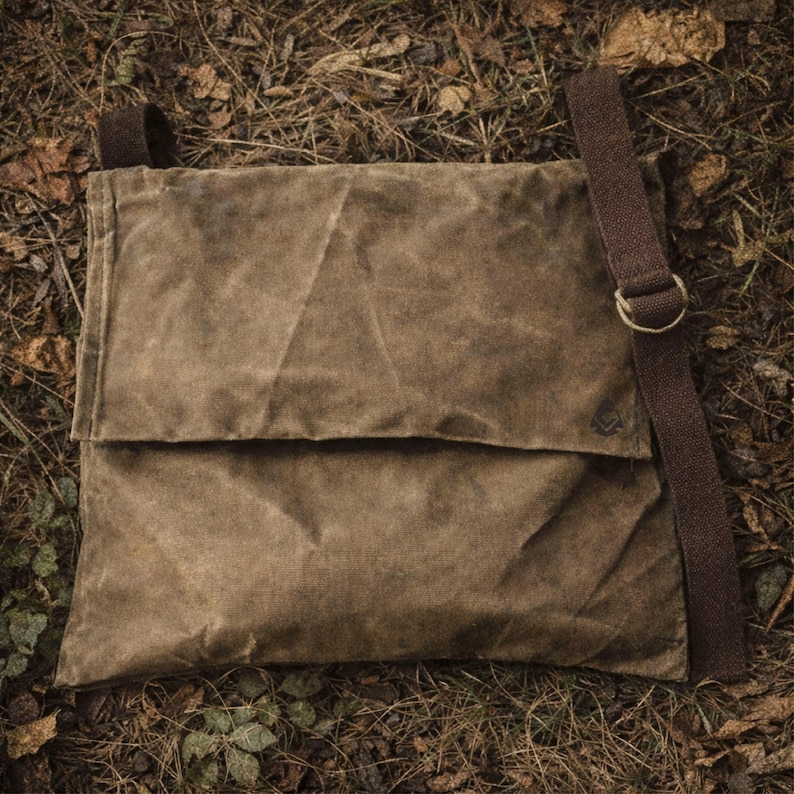 May include: A brown waxed canvas messenger bag with a fold-over flap closure. The bag features a dark brown adjustable fabric shoulder strap attached with a metal ring. The material has a textured, weathered appearance with visible creases.