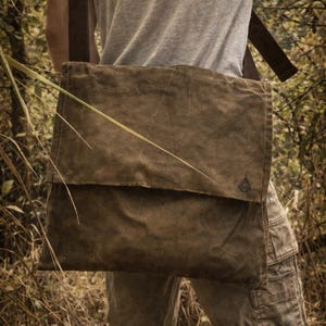 May include: A dark olive green messenger bag with a large rectangular flap and a wide shoulder strap. The bag features a textured, weathered appearance and a small, dark triangular logo emblem positioned on the lower right corner of the flap.