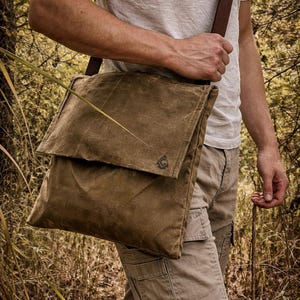 May include: A brown waxed canvas messenger bag with a fold-over flap and a dark brown adjustable shoulder strap. The bag features a small, dark triangular logo embossed on the lower right corner of the flap, providing a structured look.