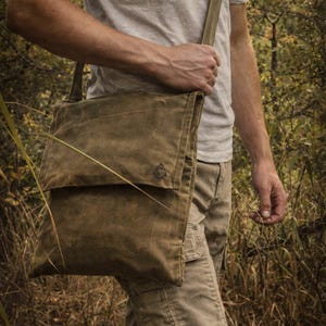 May include: A rectangular olive green waxed canvas messenger bag with a fold-over flap, an adjustable shoulder strap with a metal D-ring, and a small triangular logo embossed on the lower right corner of the front flap.