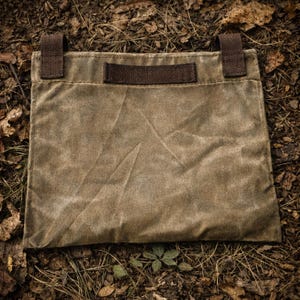 May include: A rectangular olive green waxed canvas pouch featuring a dark brown fabric handle centered on the top edge and two dark brown fabric loops attached to the upper corners. The pouch has a visible textured surface with creases.