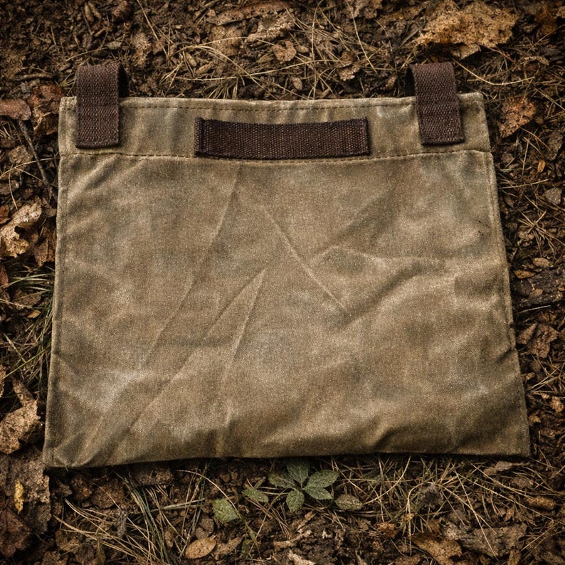 May include: A rectangular olive green waxed canvas pouch featuring a dark brown fabric handle centered on the top edge and two dark brown fabric loops attached to the upper corners. The pouch has a visible textured surface with creases.