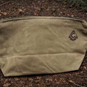 May include: A tan-colored zippered pouch made of a textured fabric. The pouch features a dark brown logo on the upper right side, which depicts a stylized arrowhead shape containing a geometric arrow symbol pointing toward the upper right corner.