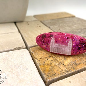 May include: Pink oval-shaped resin hair clip with a white glitter embedded image of a building. The clip has pink glitter embedded in the resin.