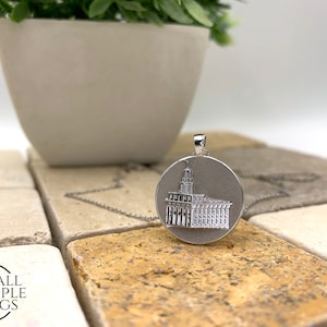 May include: Silver pendant necklace featuring a detailed engraved image of a building. The pendant is round and hangs from a silver chain.