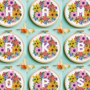 May include: Ten embroidery hoops with floral designs and letters spelling out "H A M I L T O N" in white. The hoops are arranged in two rows of five, on a light blue background.