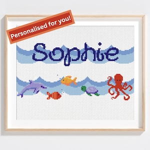 May include: Cross-stitch artwork featuring the name "Sophie" in blue, above a scene of ocean waves and sea creatures. Includes a dolphin, fish, turtle, and octopus. The words "Personalised for you!" are on a red banner.