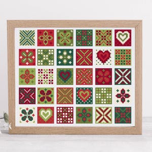 May include: A framed cross-stitch pattern featuring a grid of squares with various designs. The squares are primarily red, green, and white, with patterns including hearts, flowers, and geometric shapes. The frame is made of light-colored wood.