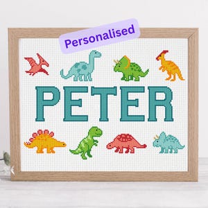May include: Cross-stitch artwork in a wooden frame featuring the name "PETER" in teal block letters. The design includes various colourful dinosaur illustrations. A banner at the top reads "Personalised".