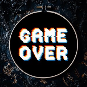 May include: A black cross-stitch hoop with the words "GAME OVER" in pixelated white, red, and blue letters. The design has a retro video game aesthetic. The hoop is set against a dark, textured background.