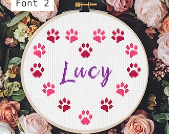 May include: A white embroidery hoop with a purple cursive name "Lucy" surrounded by pink and red paw prints.