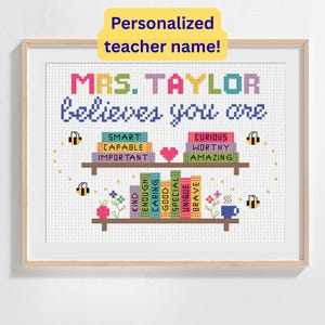 May include: A framed cross-stitch artwork with the text "MRS. TAYLOR believes you are" above two shelves of books. The books have words like "Smart," "Capable," and "Curious." The artwork is personalized with a teacher's name.