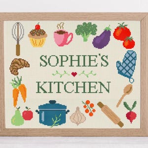 May include: Cross-stitch artwork in a wooden frame with a kitchen theme. The design includes food items such as a cupcake, tomatoes, and a pot, alongside the text "SOPHIE'S KITCHEN" in green.