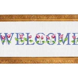 Könnte beinhalten: Ein gerahmtes Kreuzstich-Kunstwerk mit dem Wort "WELCOME" in blauen Buchstaben, verziert mit rosa Blumenmotiven. Der Rahmen ist goldfarben und verleiht dem handgefertigten Design eine elegante Note.