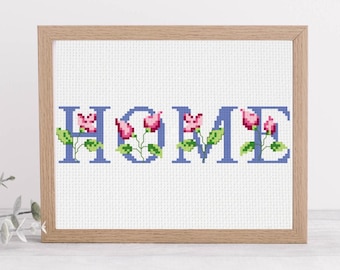 May include: Cross-stitch artwork in a wooden frame. The word "HOME" is stitched in blue, with pink flowers and green leaves incorporated into the letters. The background is white.
