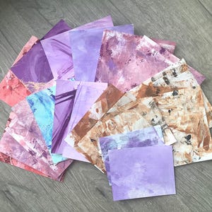 May include: A collection of square art papers in various shades of purple, pink, and brown. The papers have abstract painted designs with visible brushstrokes and textures. The papers are arranged in a fan shape, showcasing the different color variations.