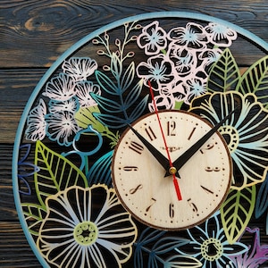 K&ouml;nnte beinhalten: Eine runde Holzuhr mit floralem Design. Das Zifferblatt ist hell mit schwarzen Zeigern und einem roten Sekundenzeiger. Der &auml;u&szlig;ere Ring zeigt lasergeschnittene Blumen und Bl&auml;tter in Blau-, Gr&uuml;n- und Rosat&ouml;nen. Die Uhr befindet sich vor einem dunklen Holzgrund.