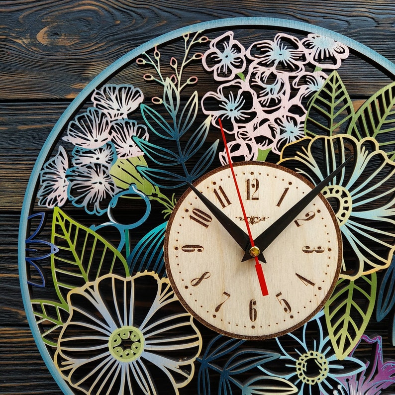K&ouml;nnte beinhalten: Eine runde Holzuhr mit floralem Design. Das Zifferblatt ist hell mit schwarzen Zeigern und einem roten Sekundenzeiger. Der &auml;u&szlig;ere Ring zeigt lasergeschnittene Blumen und Bl&auml;tter in Blau-, Gr&uuml;n- und Rosat&ouml;nen. Die Uhr befindet sich vor einem dunklen Holzgrund.
