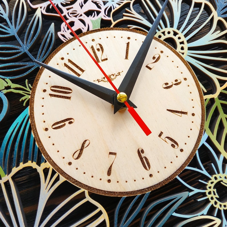 K&ouml;nnte beinhalten: Eine Wanduhr aus Holz mit hellem Zifferblatt und schwarzen Zeigern. Das Zifferblatt hat braune Ziffern und einen roten Sekundenzeiger. Die Uhr ist von einem lasergeschnittenen Blumendesign in Blau-, Gr&uuml;n- und Rosat&ouml;nen umgeben.
