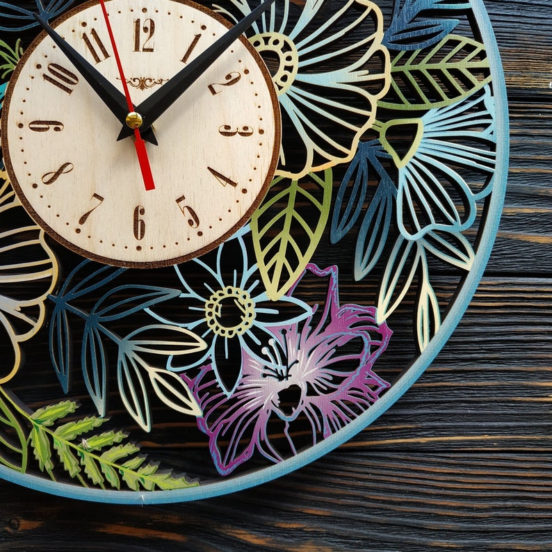 K&ouml;nnte beinhalten: Eine dekorative Holzuhr mit floralem Design. Das Zifferblatt ist hell mit schwarzen Ziffern und Zeigern. Der &auml;u&szlig;ere Ring zeigt lasergeschnittene Blumen in Blau-, Gr&uuml;n- und Lilat&ouml;nen mit einem hellblauen Rand. Die Uhr befindet sich vor einem dunklen Holzhintergrund.