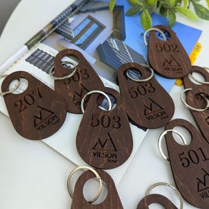 May include: A selection of wooden key fobs with silver rings. Each fob is a dark brown colour and features engraved numbers and the "VILSON hotel" logo. The numbers include 207, 501, 502, and 503.