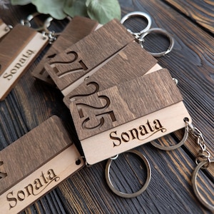 May include: Wooden keyrings with a rectangular design. The keyrings have a light wood base with a darker wood top. Some keyrings have the number "25" or "27" cut out, while others have the name "Sonata" engraved.