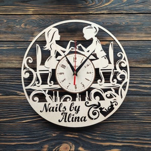 Può includere: Un orologio in legno con sfondo bianco e lancette nere. Il quadrante dell'orologio è circondato da un design decorativo di due donne che si fanno le unghie. L'orologio ha il testo "Nails by Alina" in basso.