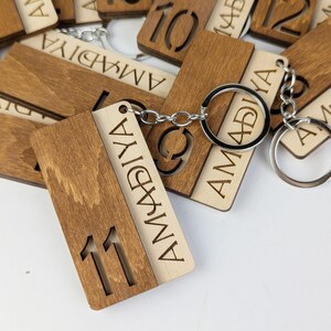May include: Wooden keyrings with engraved numbers and the name "AMADIYA". The keyrings are made of light and dark brown wood, with silver key rings. The number "11" is cut out on one keyring.
