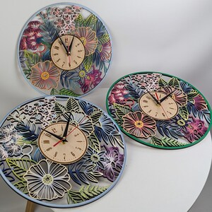 May include: Three decorative wall clocks with floral designs. Each clock features a light-colored clock face with black hands and numbers. The clocks are surrounded by laser-cut flowers and leaves in shades of pink, purple, blue, and green.