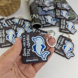 May include: Wooden keyrings with a blue and white dolphin design. The keyrings have a dark wood grain background with Ukrainian text. Each keyring has a silver key ring attached. The text reads "Day of the city with TRC DAFI, Dnipro 2025."