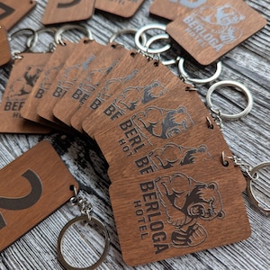 May include: Wooden keyrings with silver key rings. The keyrings are rectangular with rounded corners and feature a bear graphic and the text "BERLOGA HOTEL". The wood has a warm brown tone.