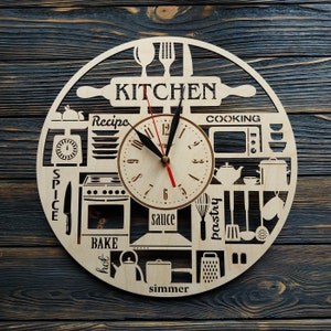 Puede incluir: Un reloj de cocina de madera con una esfera blanca y agujas negras. El reloj está rodeado de recortes de artículos de cocina, incluyendo un rodillo, una estufa, un batidor y una tetera. Las palabras "Cocina", "Especias", "Hornear", "Caliente", "Hervir a fuego lento", "Salsa", "Pastelería", "Cocinar" y "Receta" están escritas alrededor de la esfera del reloj.