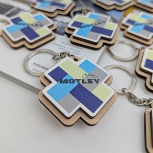 May include: Wooden keyrings with a geometric design in shades of blue, green, grey, and white. The keyrings have a silver keyring and the word "MOTLEY" in a handwritten font.