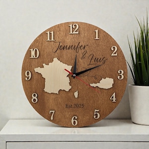 Puede incluir: Un reloj redondo de madera con acabado marrón claro. La esfera del reloj presenta un mapa de Francia y otro país, con los nombres "Jennifer & Luis" y el año "Est.2025" grabados. Las manecillas del reloj son negras y rojas.