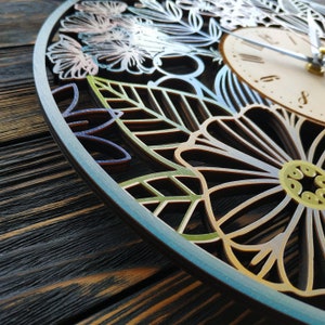 K&ouml;nnte beinhalten: Eine dekorative Holz-Uhr mit floralem Design. Das Zifferblatt ist hell mit schwarzen Ziffern und Zeigern. Der &auml;u&szlig;ere Rahmen weist aufwendige ausgeschnittene Blumen- und Bl&auml;ttermuster in Blau-, Gr&uuml;n- und Goldt&ouml;nen auf.