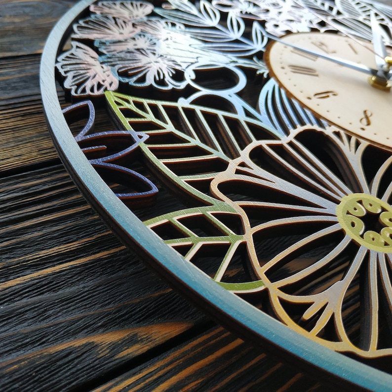 K&ouml;nnte beinhalten: Eine dekorative Holz-Uhr mit floralem Design. Das Zifferblatt ist hell mit schwarzen Ziffern und Zeigern. Der &auml;u&szlig;ere Rahmen weist aufwendige ausgeschnittene Blumen- und Bl&auml;ttermuster in Blau-, Gr&uuml;n- und Goldt&ouml;nen auf.